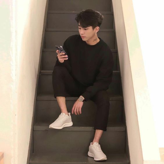 korean guy wearing an all black outfit with white shoes and sitting on stairs while using his phone