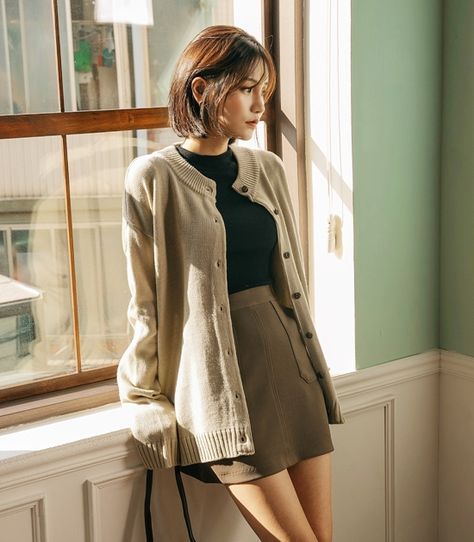 short-haired asian woman wearing a cardigan and skirt leaning against a window