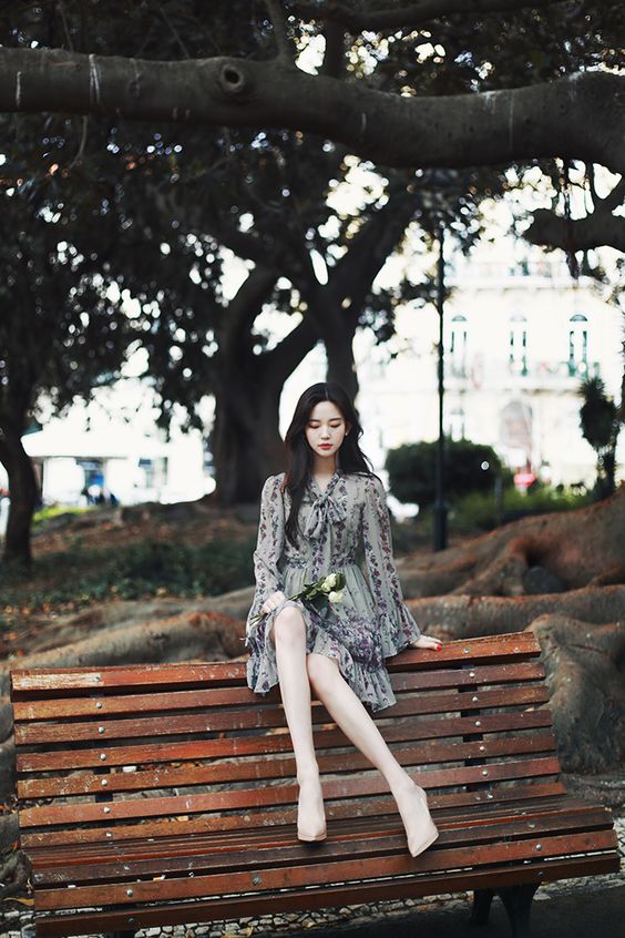 asian woman at a park wearing a long sleeved black dress and sitting on a wooden bench at the backrest with feet on the seats