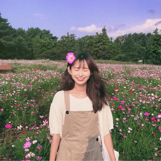 asian girl smiling with teeth in a flowerbed field wearing a dress and white top with a flower in her hair