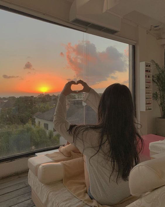 korean ulzaang girl sitting in bed and making a heart shape with her hands towards the sun