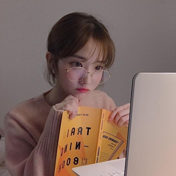 asian girl wearing glasses and looking at laptop while holding a book