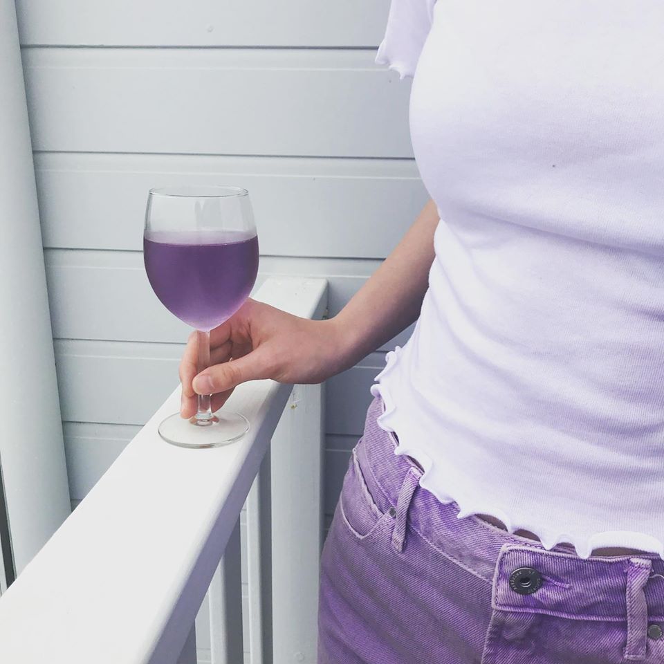 woman wearing purple top and jeans holding a glass of purple coloured Purple Reign